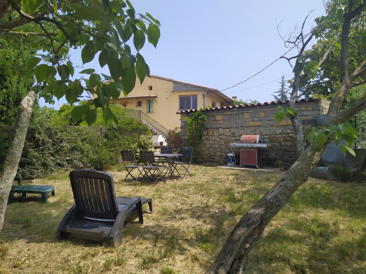 A spacious outdoor area features a grassy setting with sun loungers and a table with chairs. A barbecue grill is positioned nearby, while a stone wall and greenery provide a natural backdrop. The gîte is visible in the background, showcasing its rustic charm.