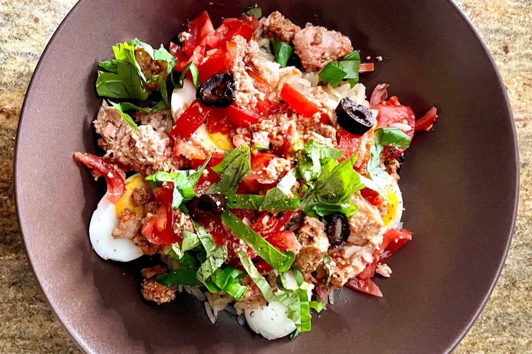 A bowl filled with a colorful mix of herbs and fresh ingredients. The dish includes chopped tomatoes, olives, herbs, and pieces of cooked meat, topped with a hint of seasoning. Soft-cooked eggs can be seen nestled among the ingredients, complementing the vibrant presentation.