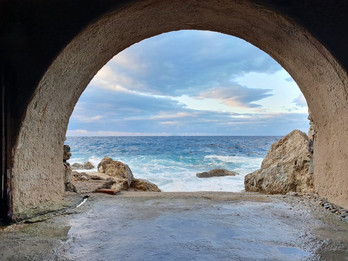 An arched opening frames a striking view of the sea, with waves crashing against rocks. The sky features soft clouds and varying shades of blue, inviting a sense of tranquility. The ground appears wet, reflecting the natural beauty of the coastal setting.