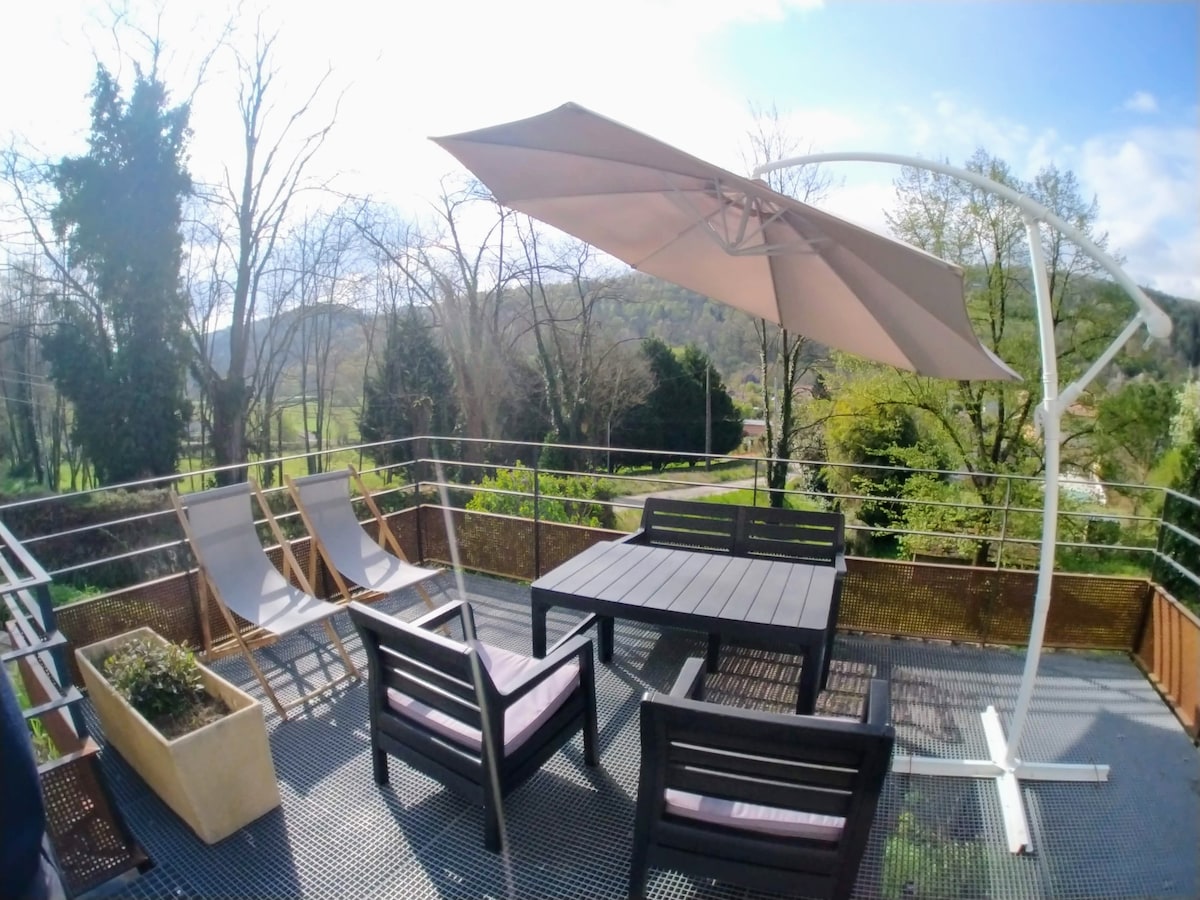 A spacious terrace is shown, featuring a large table surrounded by chairs. Two reclining chairs are positioned nearby under a large umbrella. The view captures lush greenery and rolling hills in the distance, creating a serene outdoor setting.