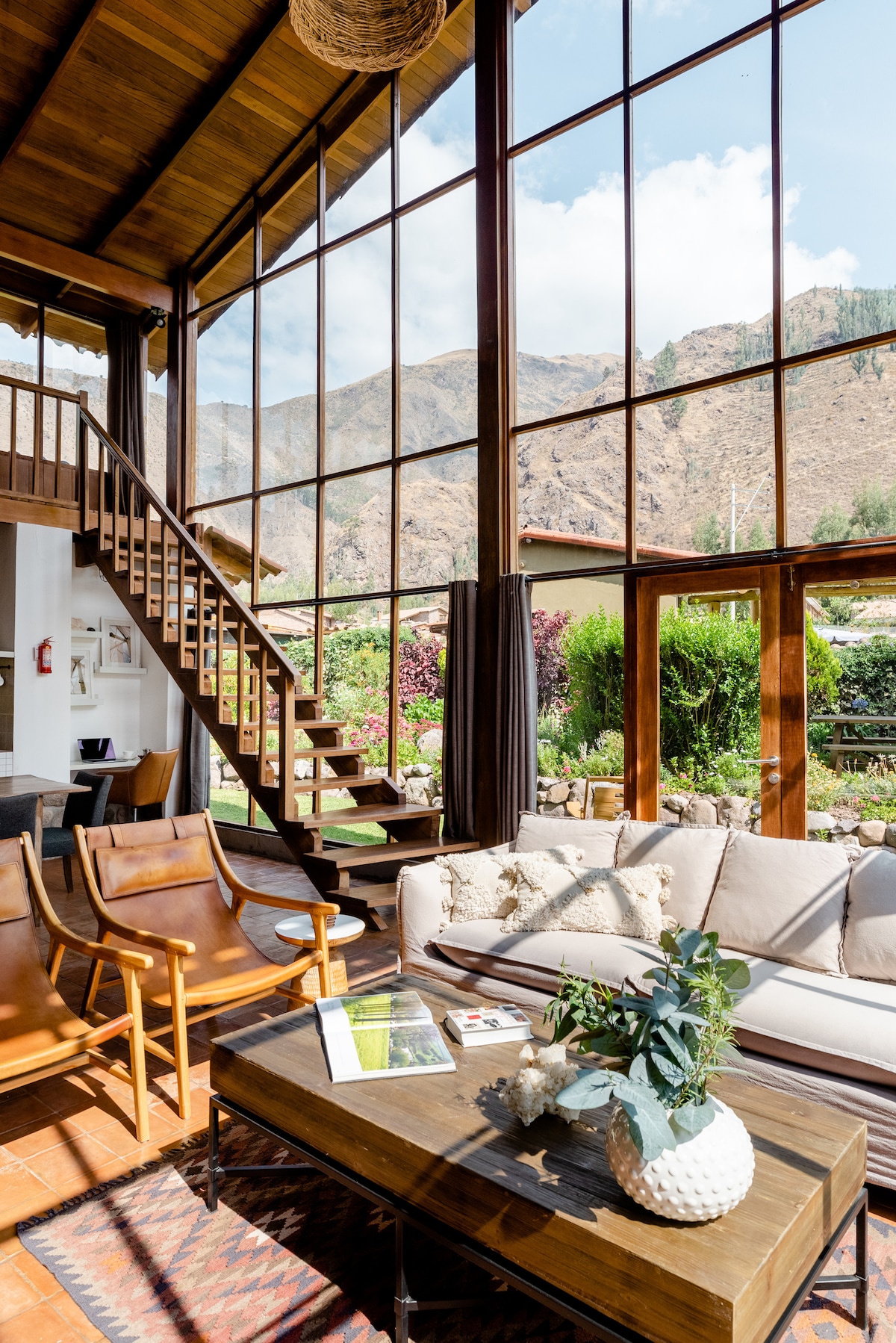 A spacious living area is highlighted by large floor-to-ceiling windows that provide views of the surrounding mountains. A comfortable sofa and a wooden coffee table occupy the space, complemented by stylish armchairs and a decorative plant. Warm wooden accents add a cozy atmosphere.