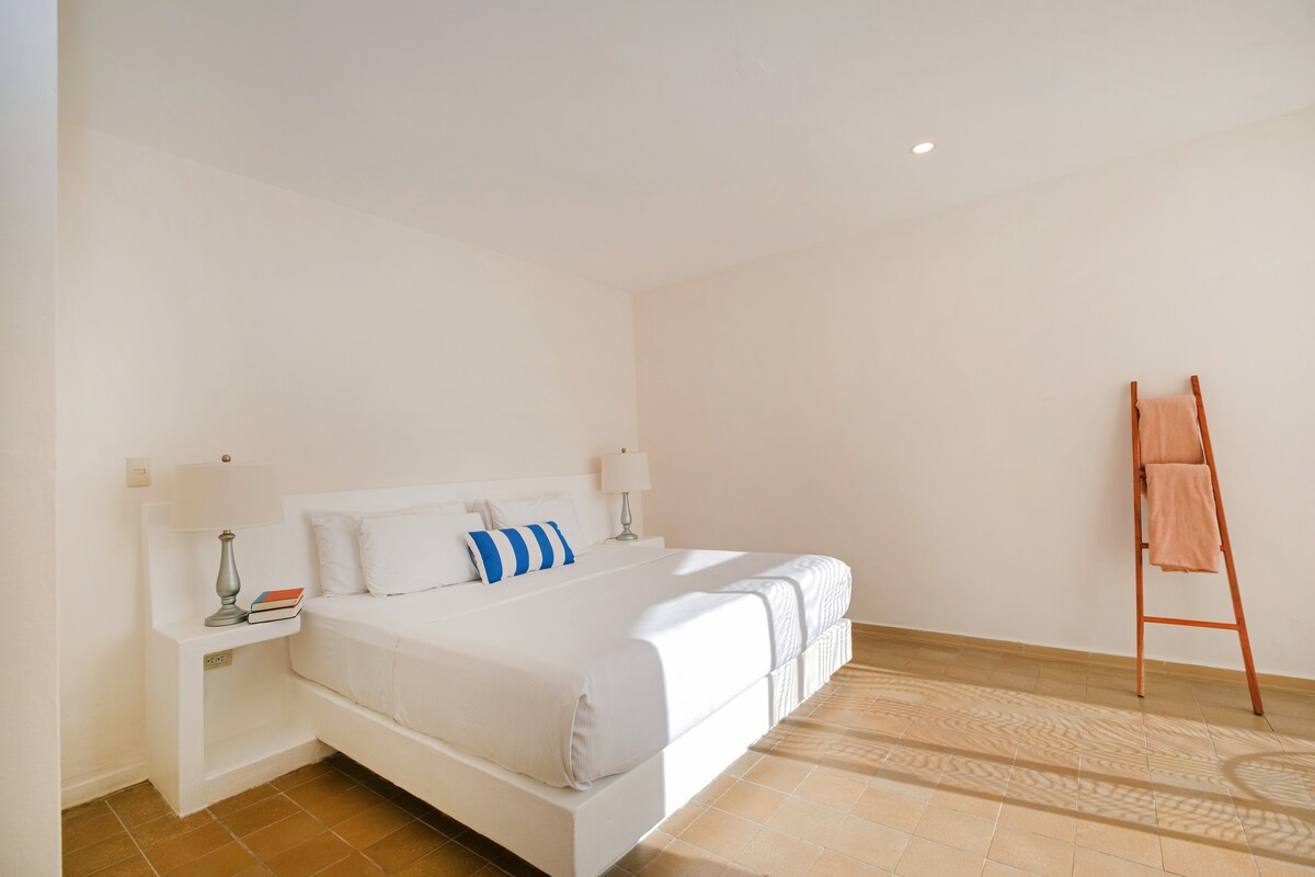 A spacious bedroom features a large bed with white linens accented by a striped decorative pillow. Two bedside lamps are positioned on either side. A wooden ladder holds a towel, and natural light fills the room through unobstructed windows, illuminating the neutral tones of the walls and flooring.