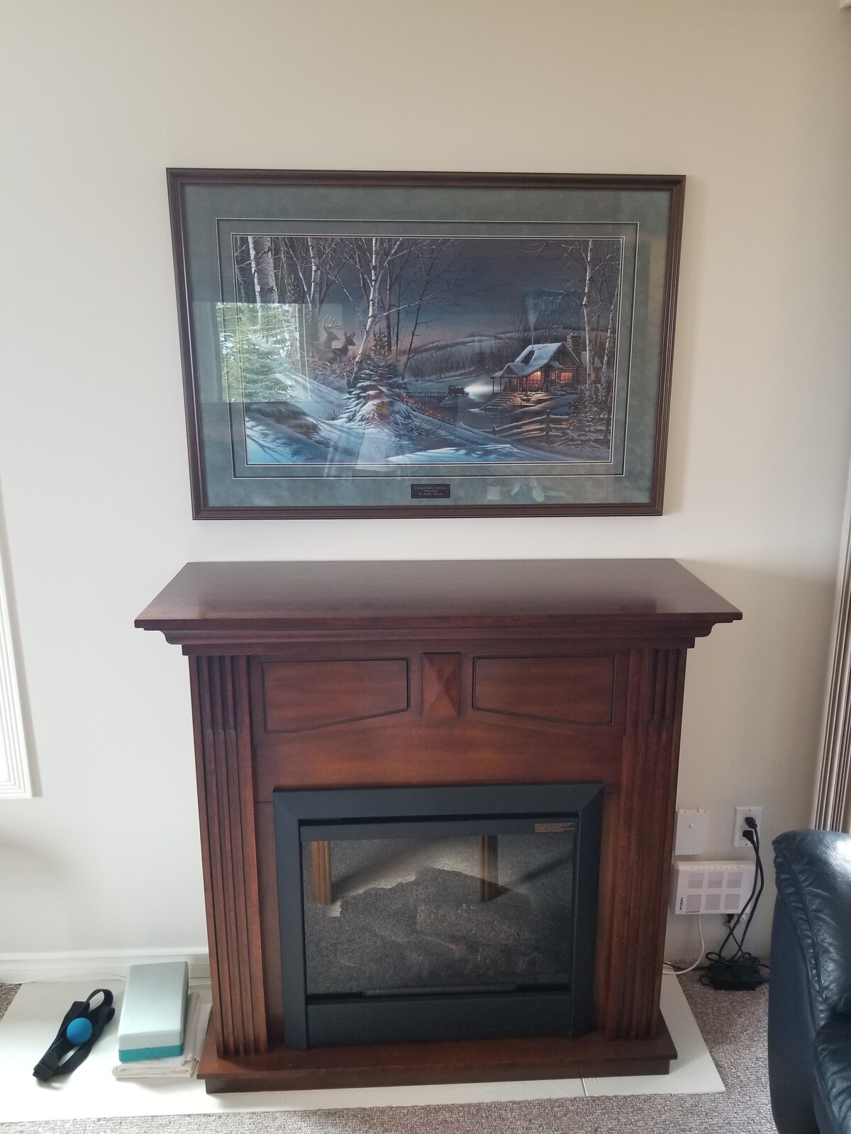A wooden fireplace mantle is positioned against a light-colored wall. Above it hangs a framed artwork depicting a winter scene with a cabin and snow-covered trees. Natural light streams in from nearby windows, casting soft shadows.