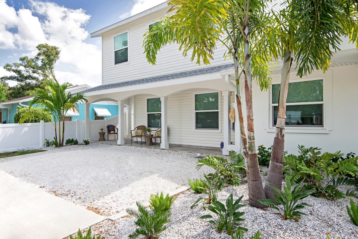 The exterior of the cottage is surrounded by a charming landscape, featuring various tropical plants and a gravel driveway. A covered porch offers seating, while the light-colored home creates an inviting appearance under the bright sky.