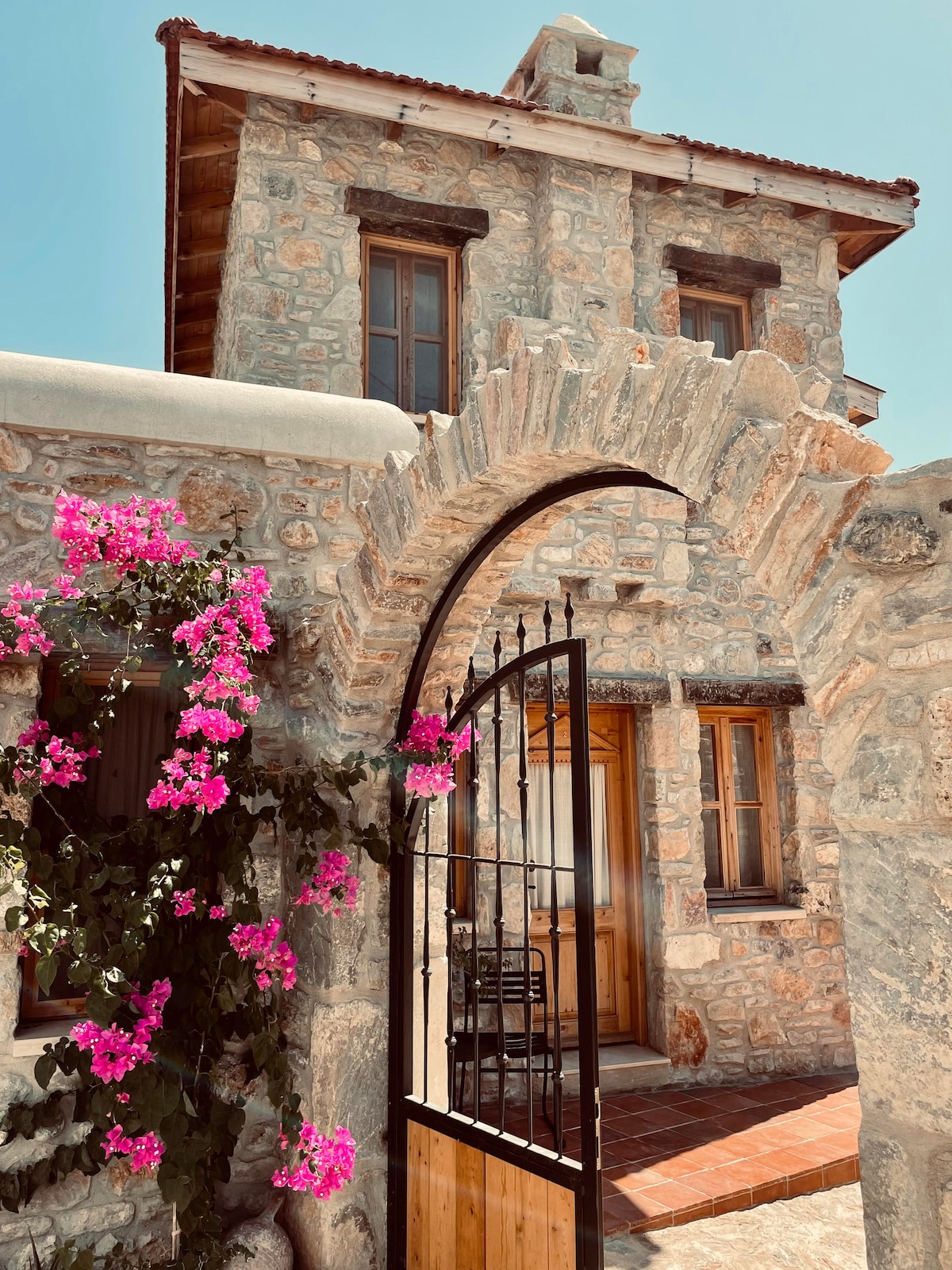 An arched stone gateway adorned with vibrant pink bougainvillea leads to a rustic stone house. Sunlight highlights the building's traditional architecture, featuring wooden windows and a warm-toned door, inviting guests to explore further.