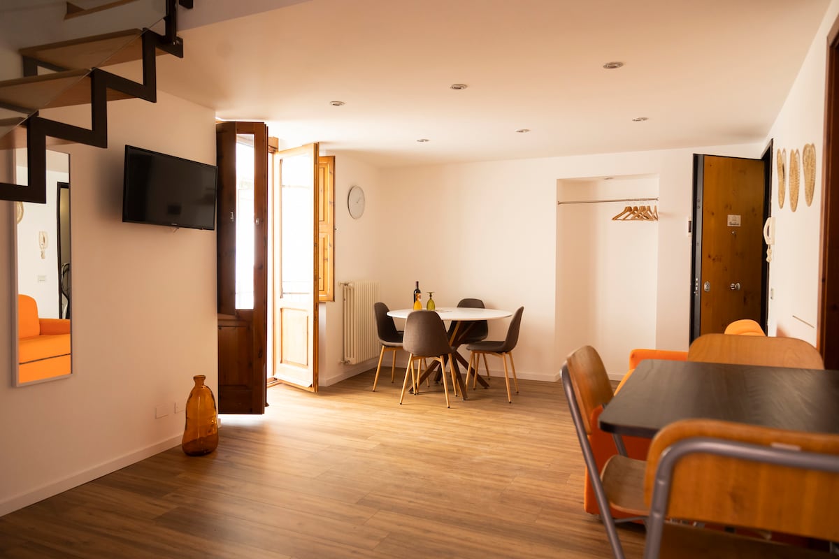 The open living area is characterized by warm wooden flooring and natural light entering through large windows. A dining table with chairs is positioned near the windows, while a cozy seating area featuring a bright sofa enhances the inviting space. A mirror reflects the room's design.