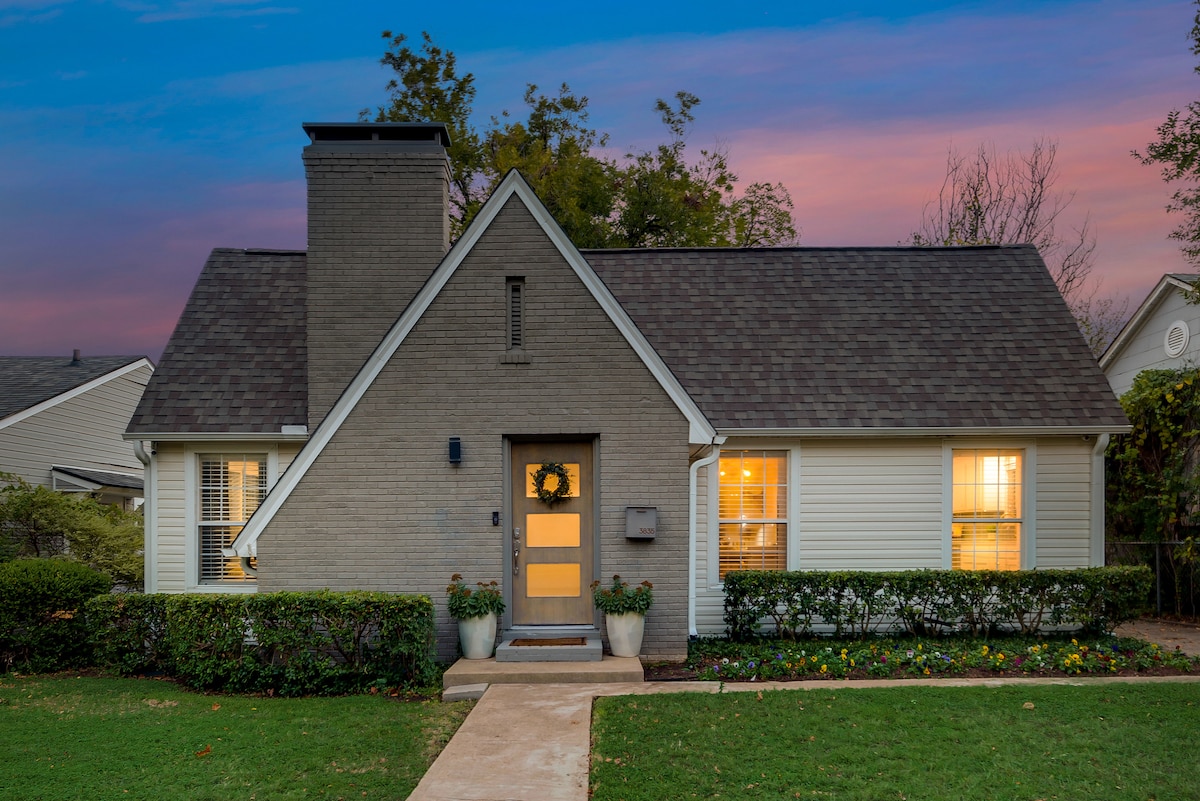 The bungalow features a classic architectural style with a brown shingled roof and a central chimney. Bright windows are framed by decorative shrubbery, while a welcoming pathway leads to the front door adorned with a seasonal wreath.