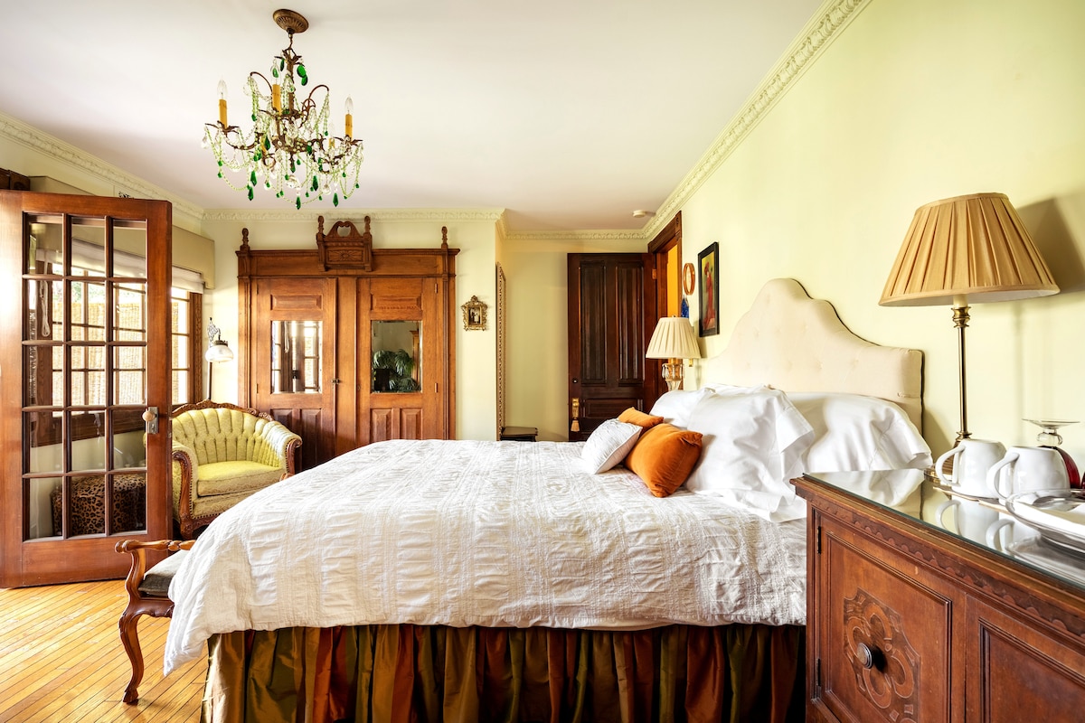 A spacious bedroom features a large bed with a white comforter and decorative pillows. An antique chandelier hangs overhead, and a vintage seating area is visible beside a wooden entrance. Elegant crown molding adorns the walls, complemented by rich wood furnishings.