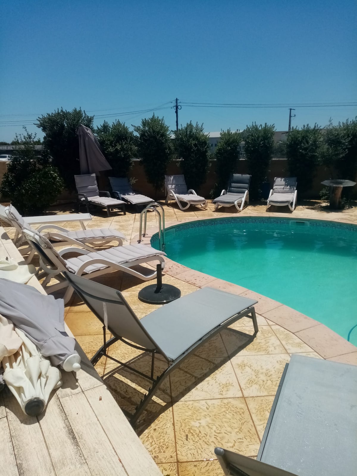 A private pool area is surrounded by tall hedges for added privacy. Sun loungers are arranged around the pool, offering a space for relaxation. A shaded umbrella is positioned beside a small table and chair set, enhancing the outdoor comfort.