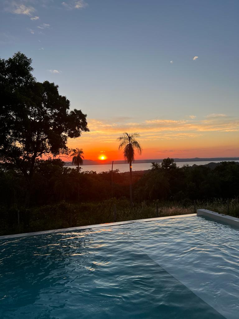 A serene sunset view is captured over a tranquil lake, framed by lush vegetation. The infinity pool reflects the warm colors of the sky, blending seamlessly with the horizon. Silhouettes of palm trees stand against the fading light, enhancing the peaceful surroundings.