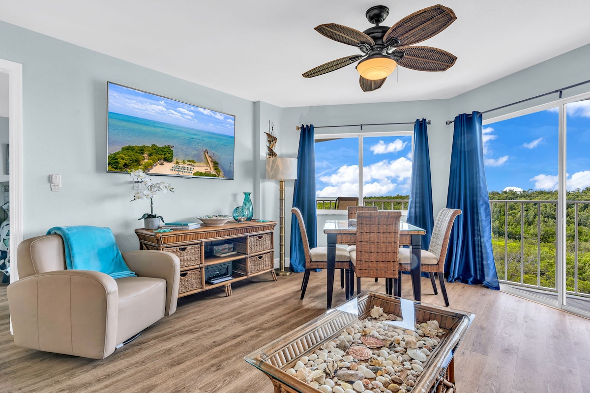 A light-filled living area features a comfortable chair and a spacious dining table. A TV is mounted on the wall, and large windows offer views of the ocean and surrounding greenery, while sheer blue curtains frame the scene.