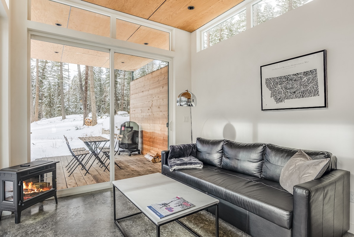 A modern living area features a sleek black sofa facing a glass sliding door that opens to a snowy outdoor deck. A compact fireplace is positioned nearby, and a minimalistic coffee table rests on a polished concrete floor. Natural light enhances the space.