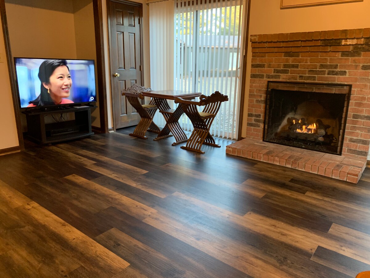 A cozy living space is presented, featuring a brick fireplace with a warm glow. A television is positioned on a stand, and a wooden table with two folding chairs is set nearby. Large windows allow natural light to brighten the room.