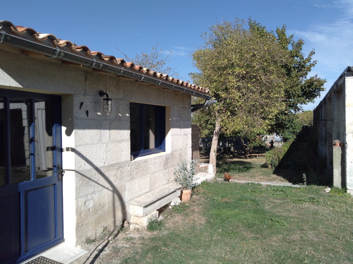 The exterior of the gîte is depicted, featuring a stone facade and a blue door. A small terrace area with a bench is visible, complemented by surrounding greenery, including a tree. The serene garden is shown, suggesting a peaceful outdoor space.