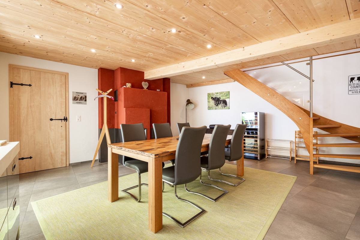 A dining area features a wooden table surrounded by six grey chairs. The space is illuminated by recessed lighting, with a wooden ceiling adding warmth. A staircase with wooden steps leads to the upper level, while a red feature wall enhances the overall design.