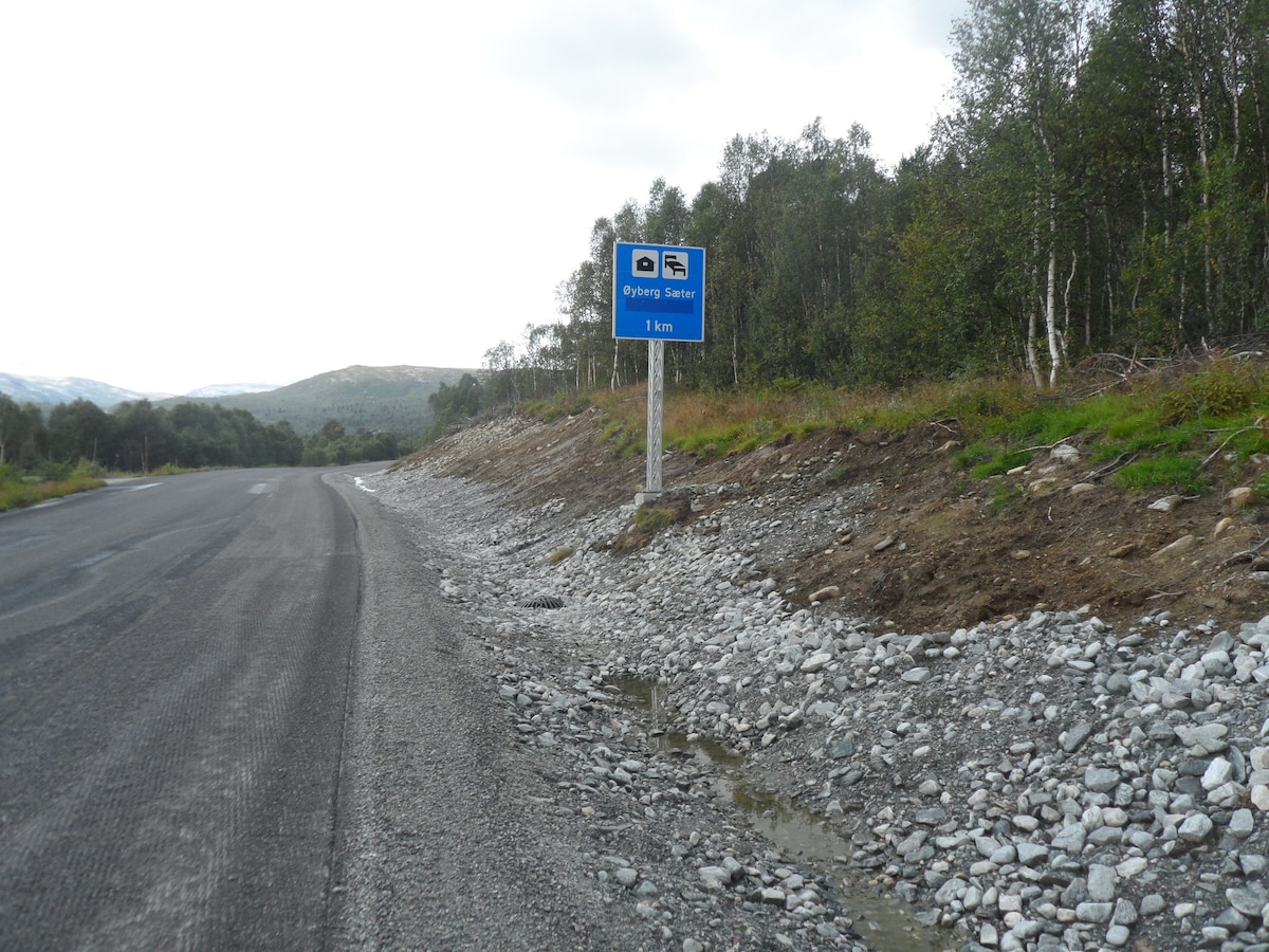 A highway sign indicating the direction to Øyberg Seter is positioned along a gravel road. The surrounding landscape features a mix of greenery, including small trees and shrubs, while the mountains are visible in the background beneath a cloudy sky.