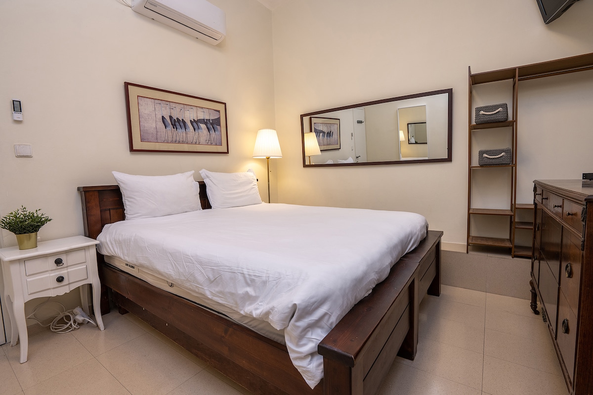 A serene bedroom is equipped with a spacious bed flanked by bedside tables. Soft lighting is provided by a lamp, and a large mirror enhances the room's sense of space. A small shelf stands in the corner, complementing the simple decor.