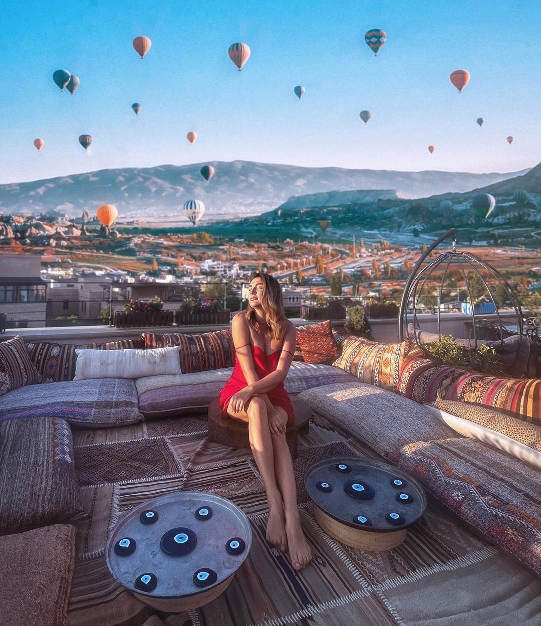 A spacious rooftop terrace is adorned with cushions and low tables, providing a comfortable seating area. In the background, colorful hot air balloons float over lush valleys and traditional village homes under a clear sky, creating a serene and picturesque view.
