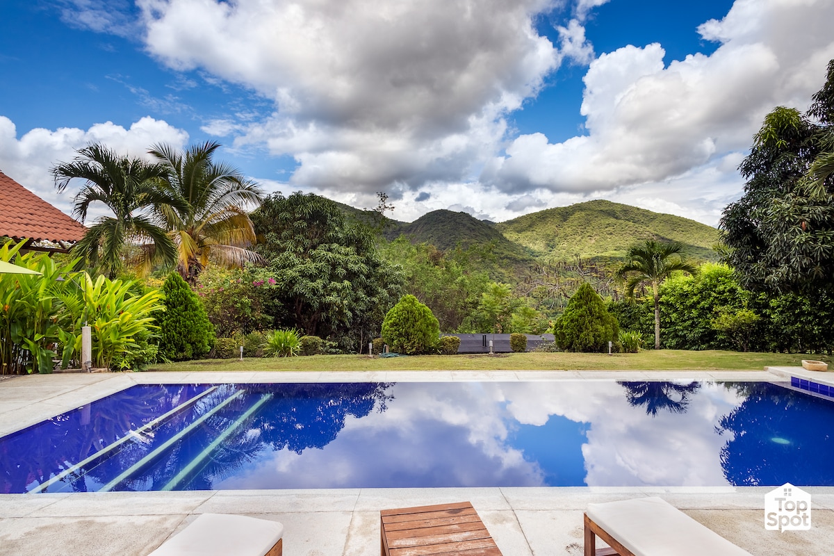 A serene private pool reflects the sky, surrounded by lush greenery and mountains. Comfortable lounge chairs are positioned nearby, inviting relaxation. The clear water glistens under soft clouds above, creating a tranquil setting for lounging and enjoying the natural beauty.