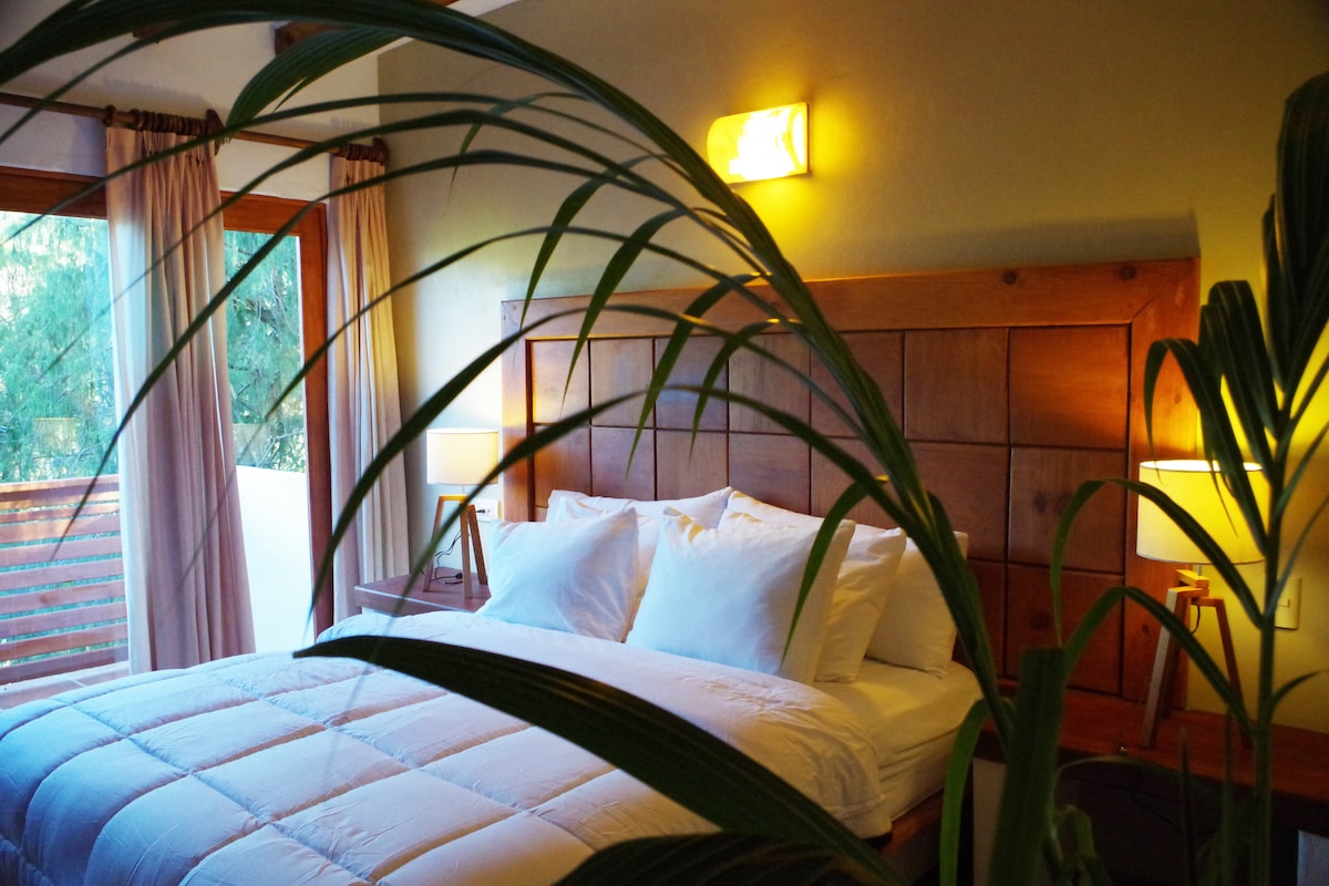 A warm-toned bedroom features a spacious bed adorned with white pillows and a light-colored comforter. Bedside lamps provide soft lighting, while large windows with sheer curtains allow natural light to fill the space. A leafy plant frames the view, adding a touch of nature.