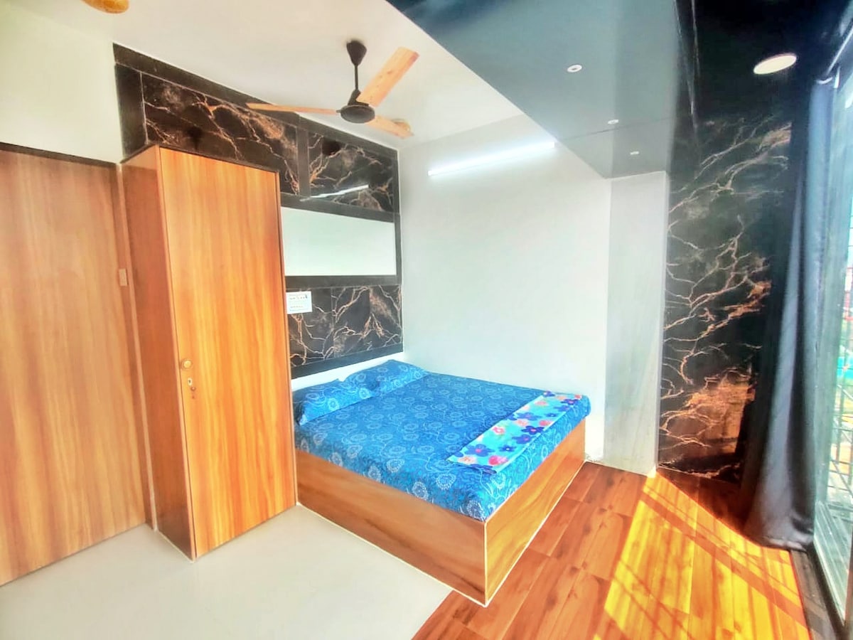 A bright bedroom is displayed, featuring a spacious bed covered with a blue patterned bedspread. A wooden wardrobe is positioned nearby, and the walls are adorned with dark marble accents. Soft, natural light streams in through a window, creating a calm atmosphere.