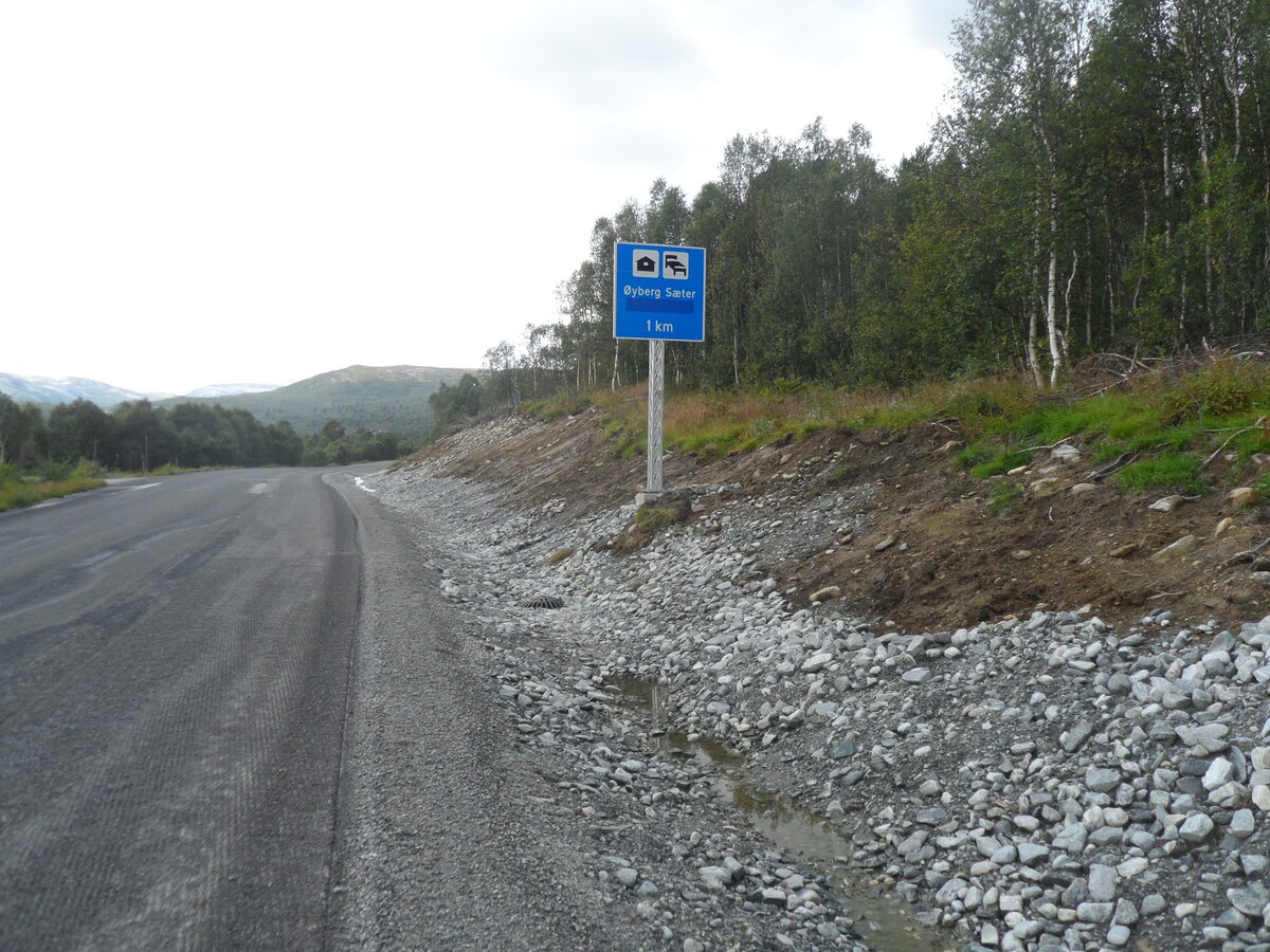 A roadside view along a well-maintained gravel path is seen, marking the approach to a destination. A blue roadside sign indicates a location 1 kilometer away, surrounded by green foliage and rocky terrain. Mountains are visible in the background under a cloudy sky.