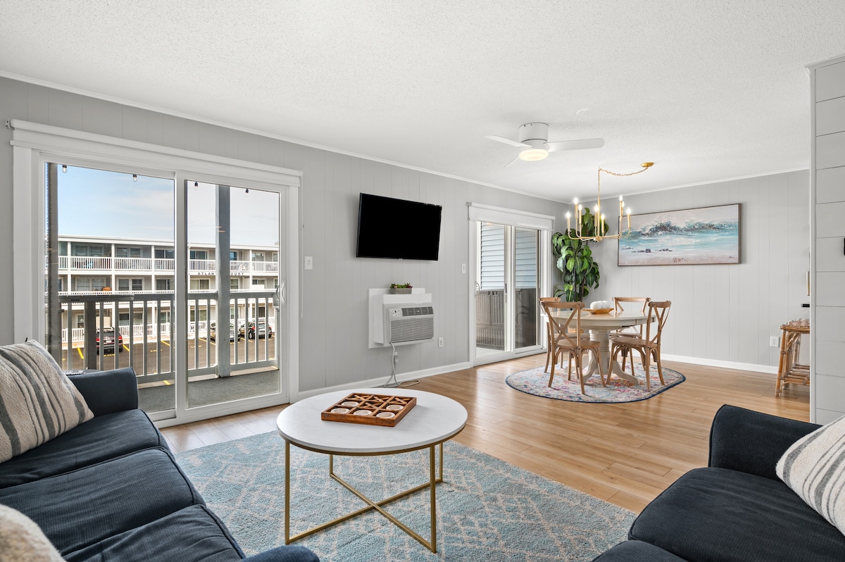 A well-lit living area showcases a comfortable sleeper sofa and a love seat, centered around a round table for four. Large glass doors provide access to a private balcony. A mounted TV is visible on the wall, adding to the functional design of the space.
