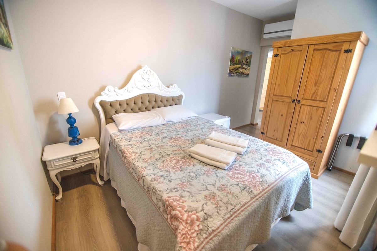 A spacious bedroom features a plush bed adorned with a floral patterned coverlet. A nightstand with a blue lamp is positioned beside the bed. A wooden wardrobe is visible in the corner, while soft natural light fills the room through the window.