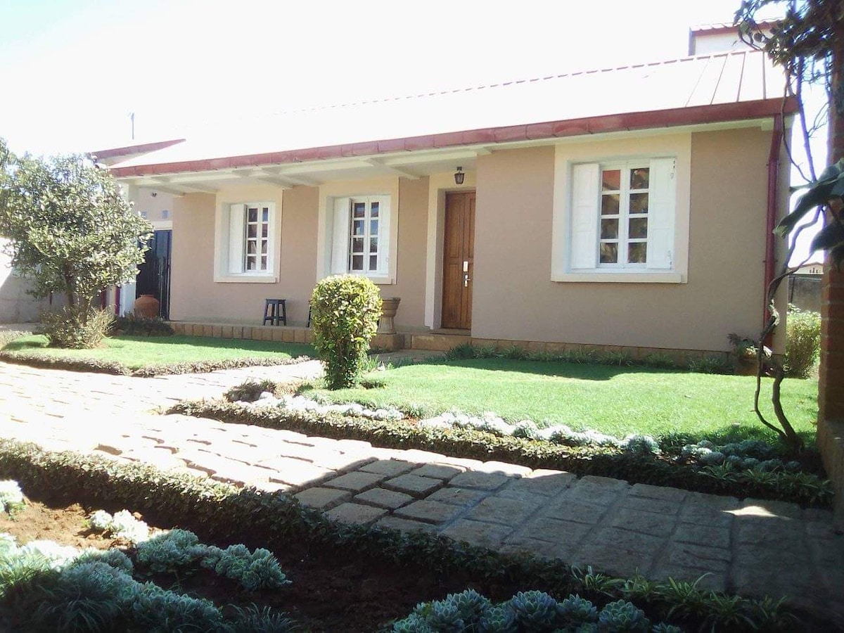 A single-story villa is presented with a well-maintained garden featuring a manicured lawn. The entrance encompasses a pathway lined with stones and lush greenery. Windows are framed with white trims, allowing natural light to fill the interior spaces. A wooden door serves as the main entry.