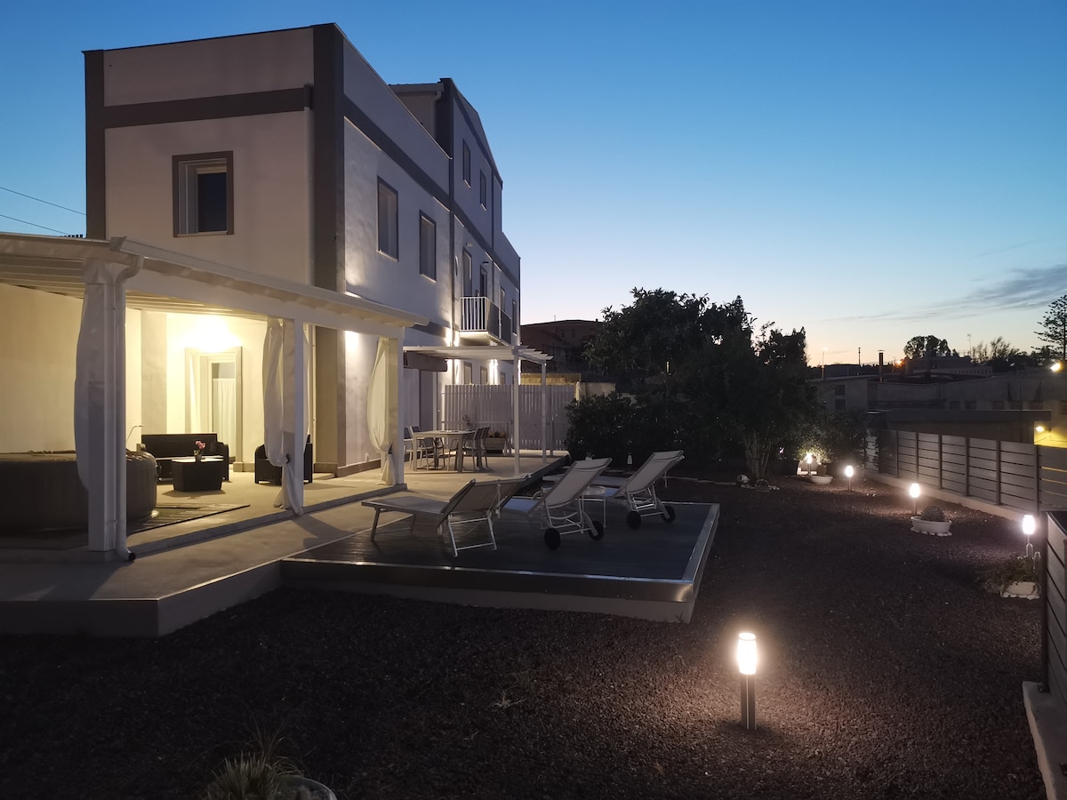 The exterior of the villa is illuminated during twilight, showcasing a spacious terrace with lounge chairs. An outdoor seating area is visible under a covered porch, while a well-kept garden features subtle lighting, enhancing the tranquil atmosphere of the space.