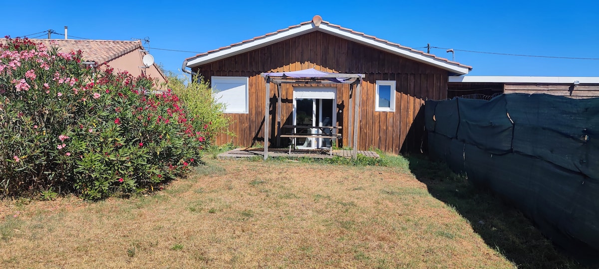 A wooden chalet features large glass doors opening to a spacious yard, surrounded by greenery and flowering shrubs. A shaded seating area is present on the deck, offering an outdoor space for relaxation. The clear blue sky enhances the inviting atmosphere of the outdoor setting.