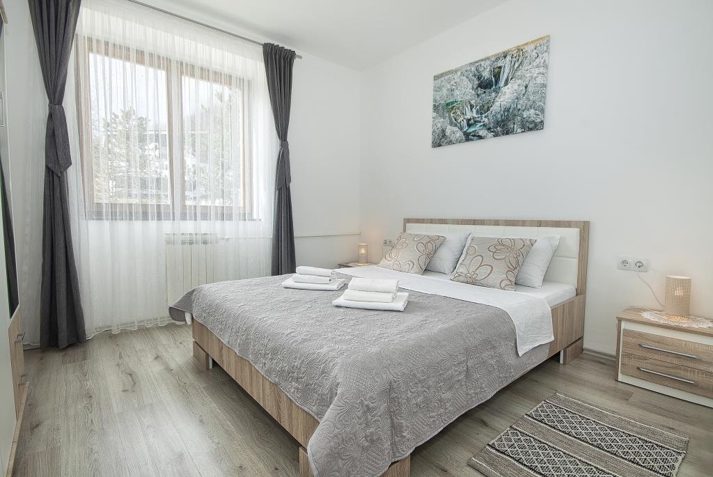 A bright bedroom features a spacious bed adorned with light bedding and pillows. Natural light filters through sheer curtains on a large window, creating a serene environment. A piece of wall art adds a subtle touch, while a small nightstand complements the room's modern decor.
