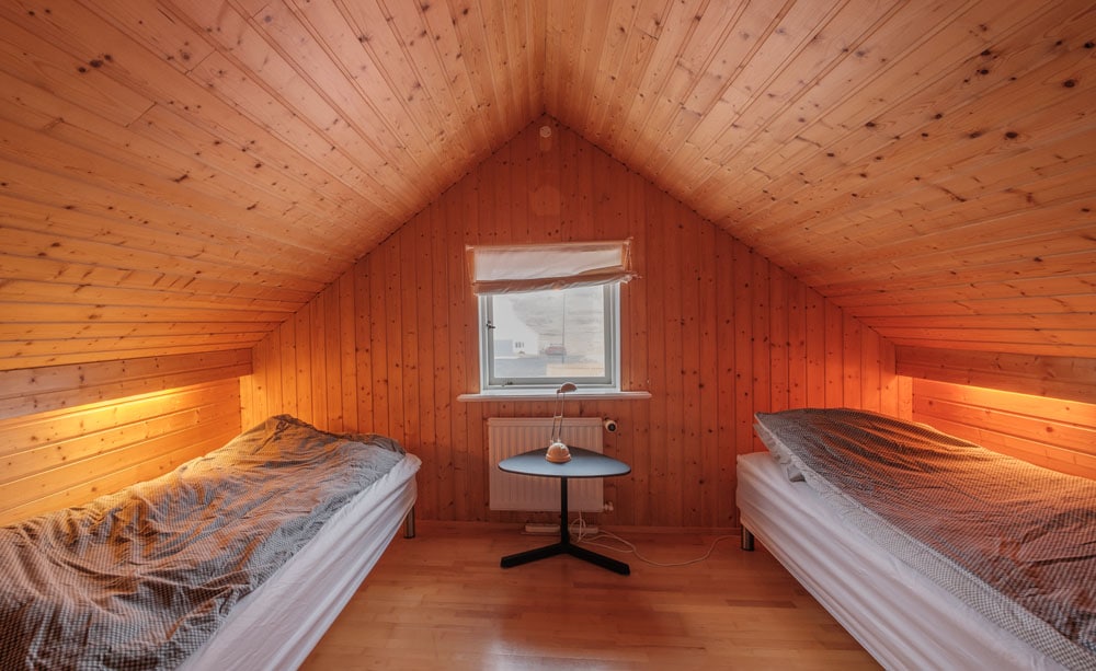 A bright, wooden-paneled bedroom is presented, featuring two twin beds with light bedding and a small table positioned between them. A window allows natural light to enter, and warm lighting is integrated along the walls, enhancing the cozy ambiance of the space.