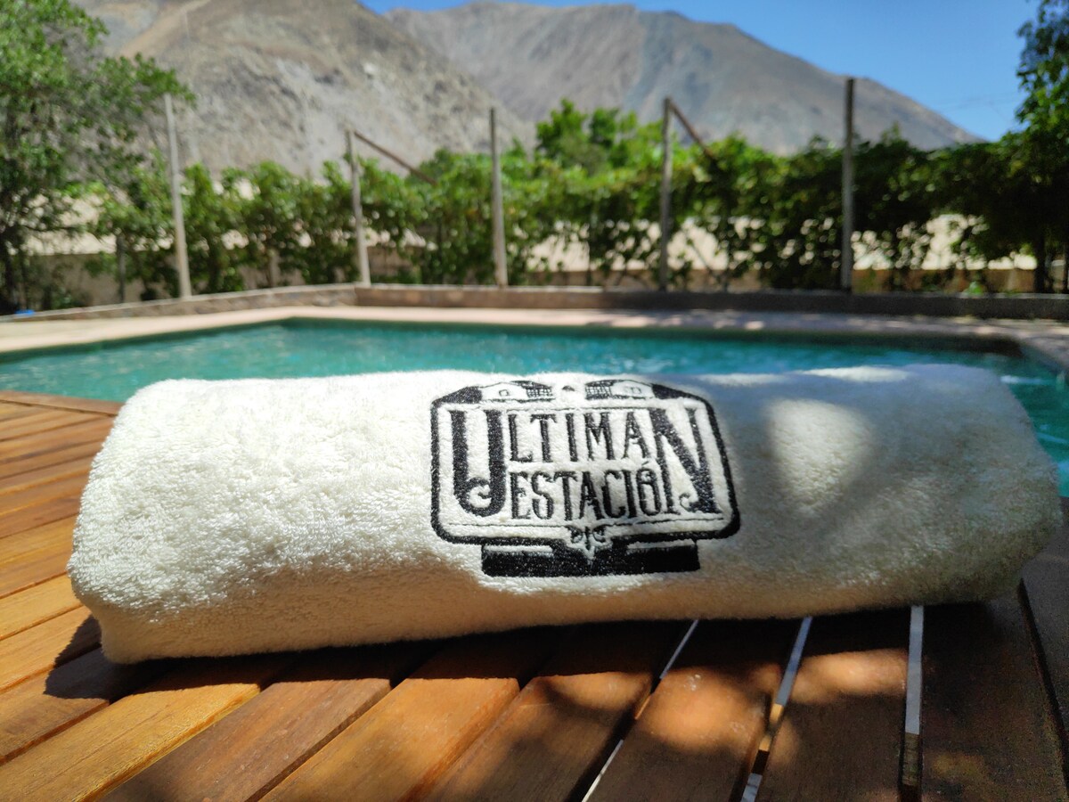 A white towel with the embroidered logo 'Ultima Estación' is neatly placed on a wooden deck. In the background, a turquoise swimming pool is visible, surrounded by green foliage and mountains, highlighting the natural beauty of the area.