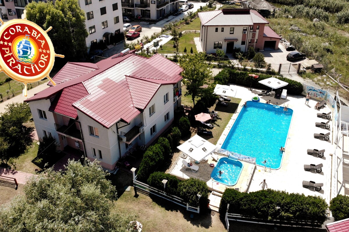 An aerial view shows the resort complex featuring a large swimming pool surrounded by lounge chairs and umbrellas. The neatly landscaped grounds include greenery and patio spaces, offering a tranquil environment. The building structure displays a modern design with a red roof.