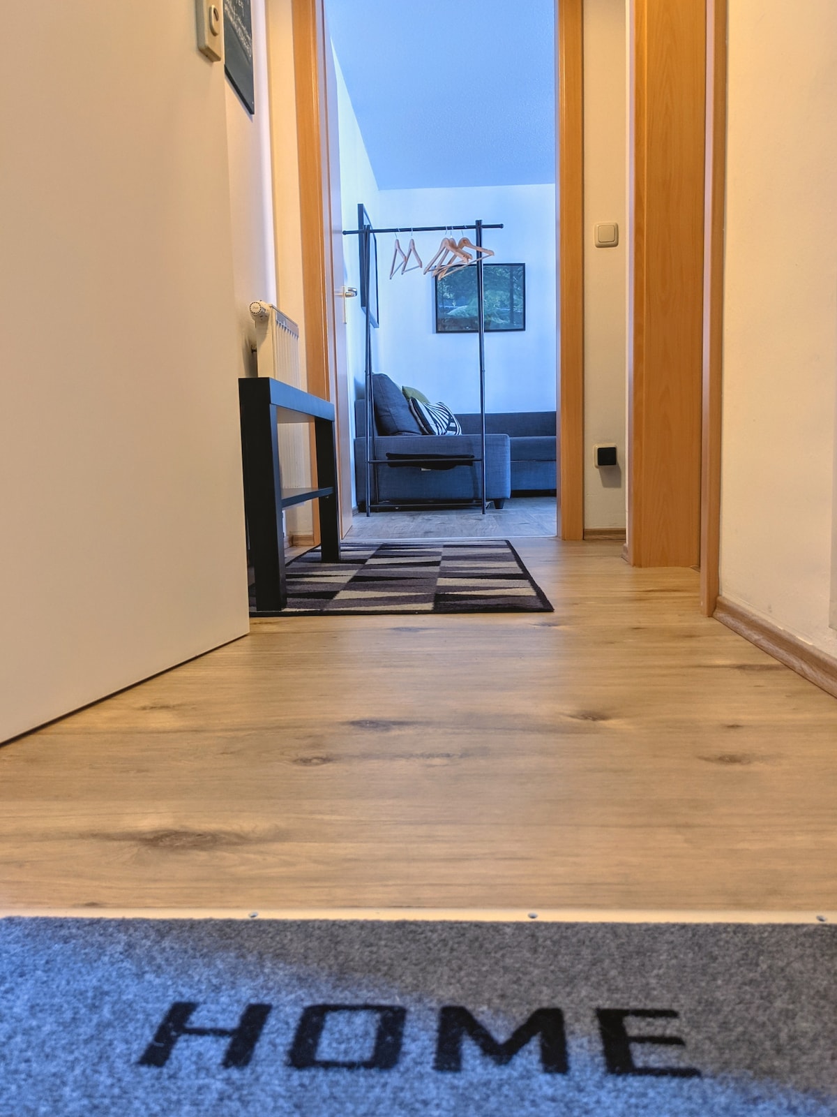 The view from the entrance captures a hallway leading to a living area, with light-colored flooring and walls. A rug with the word 'HOME' is visible at the entrance. A coat rack stands against the wall, while a cozy seating area appears in the background.