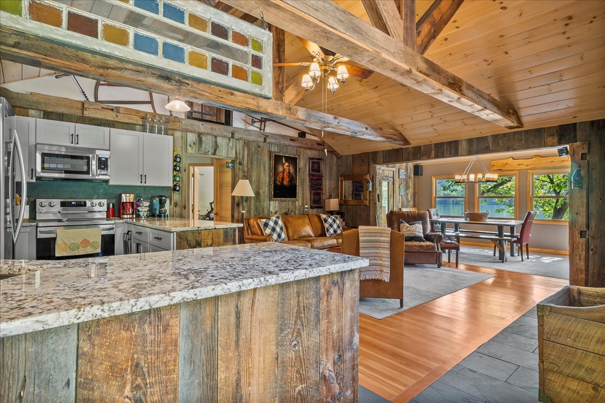 An open living space features vaulted wooden ceilings and a combination of rustic and modern decor. A large granite island offers seating, while the kitchen is equipped with stainless steel appliances. Natural light brightens the area through multiple windows, creating a welcoming environment.