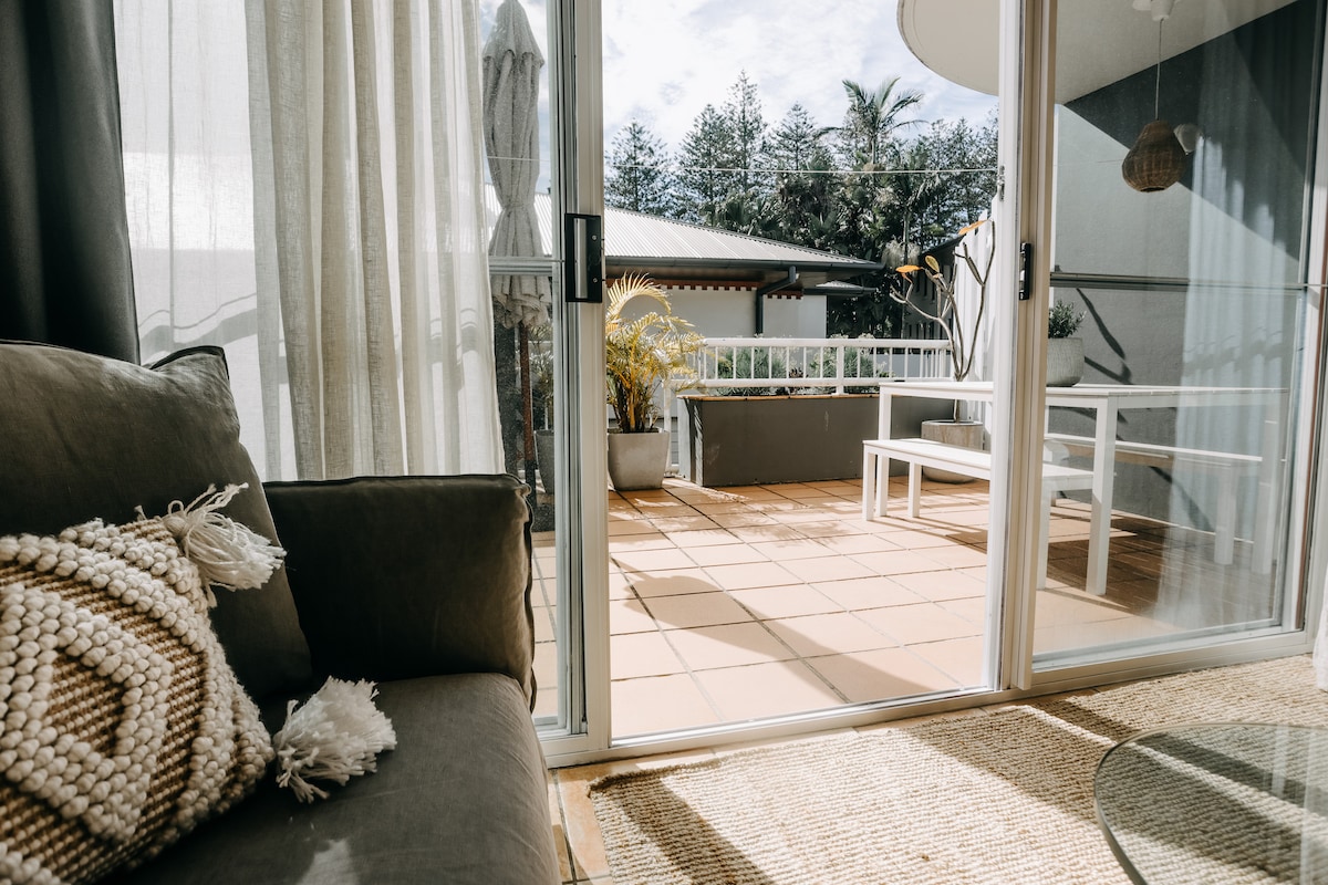 A cozy seating area features a comfortable sofa adorned with textured cushions. Light drapes softly filter sunlight, illuminating a spacious private terrace beyond, furnished with white outdoor seating and greenery, creating an inviting extension of the indoor space.