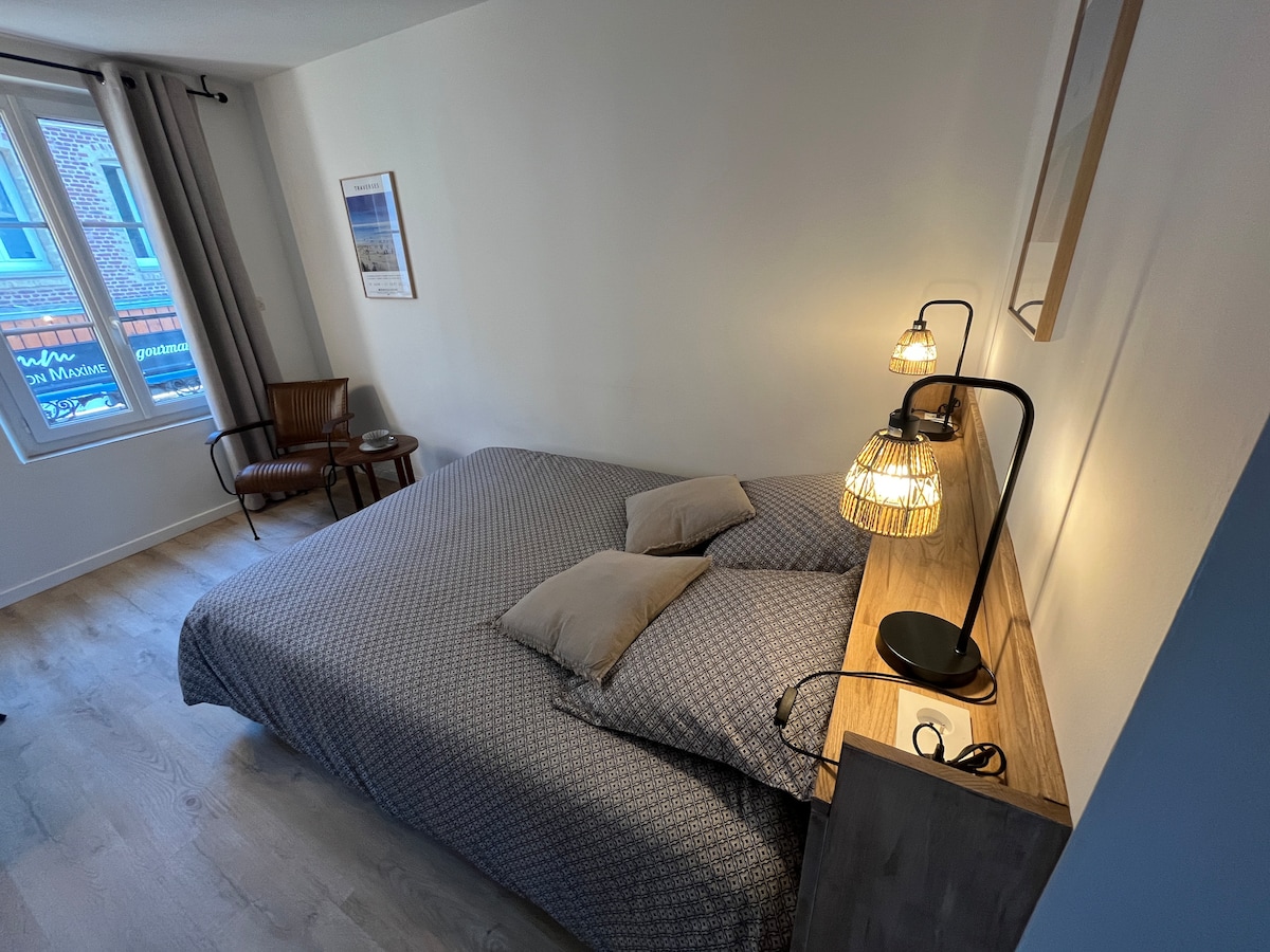 A spacious sleeping area features a large bed with patterned bedding and two pillows. Beside the bed, a wooden nightstand holds a modern lamp. Natural light enters through a window with light curtains, illuminating the light-colored walls and wooden floor.