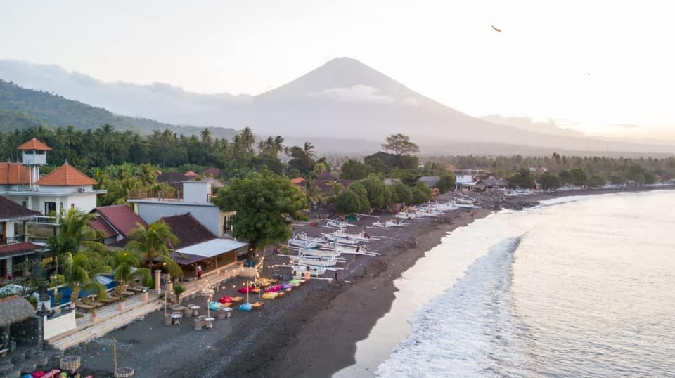 A coastal view captures a beach with a rocky shore and gentle waves lapping at the water's edge. Lush greenery and white boats dot the landscape, while the silhouette of Mt. Agung rises majestically in the background, complementing the tranquil atmosphere.