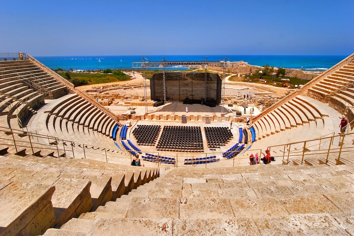 An open-air amphitheater is positioned with a view of the sea in the background. Stone seating areas are arranged in a semi-circular pattern, leading to a central stage. A clear blue sky and distant waves complete the scenic setting.