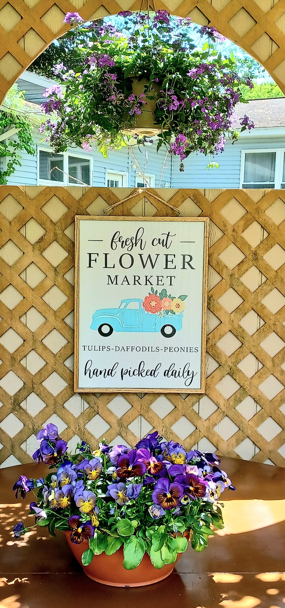 A charming flower market sign is displayed on a wooden lattice background, featuring a light blue truck and the text 'Fresh Cut Flower Market.' Below, a terracotta pot overflowing with vibrant purple and yellow pansies adds a splash of color to the scene.