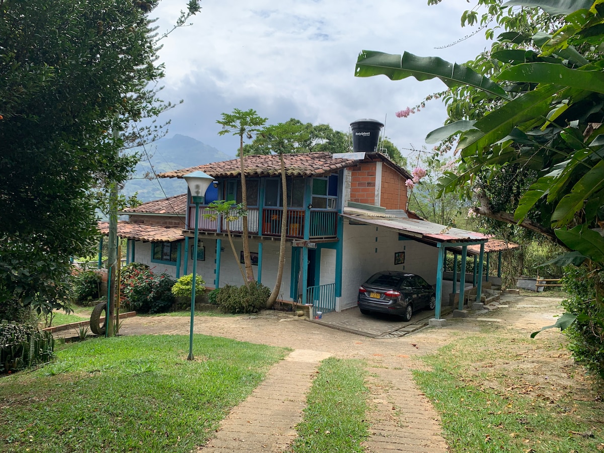 A two-story house with a blue and terracotta exterior is shown surrounded by lush greenery. A stone pathway leads to the entrance, while a parked car is visible under the shaded area. The mountains and sky are partially seen in the background.