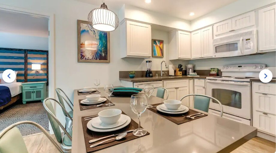 A modern kitchen and dining area are showcased, featuring a large table set for six. The countertops are complemented by white cabinetry and appliances. A glimpse of the adjacent bedroom is visible, adding a sense of openness to the space.