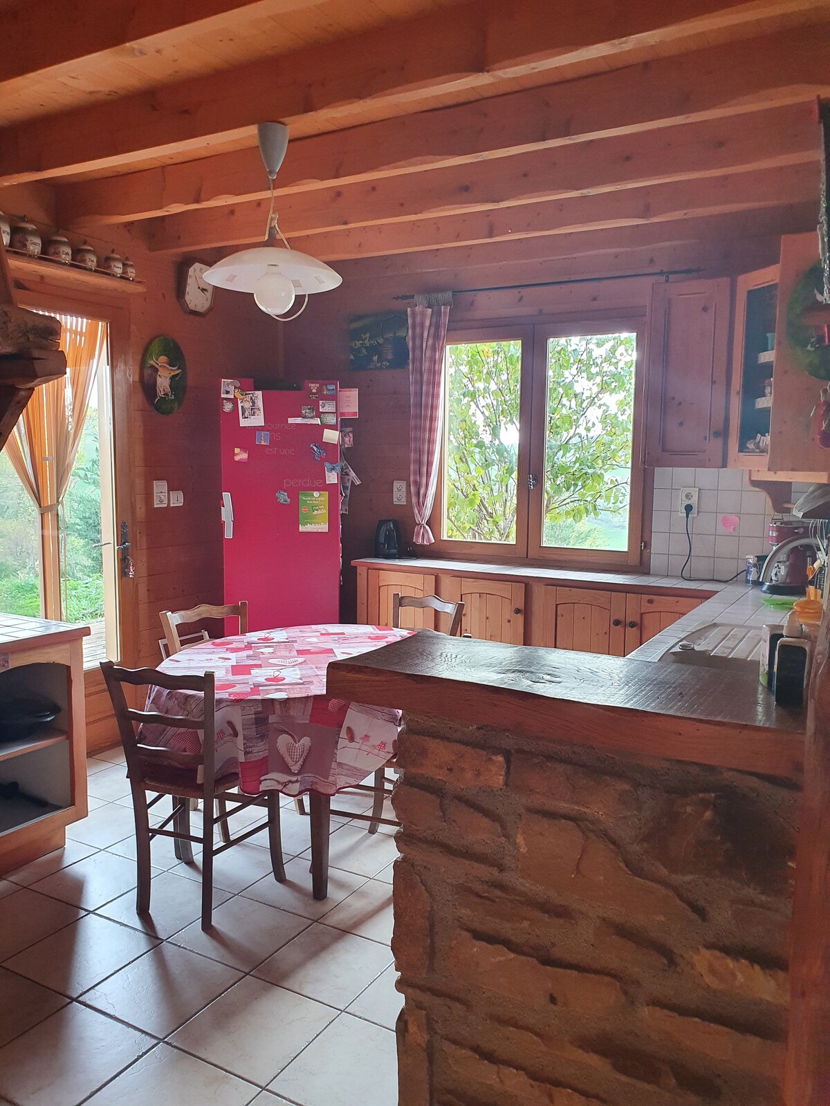 A warm wooden kitchen features rustic cabinetry and a round table set for four. A vibrant red refrigerator stands against the wall, accompanied by a window overlooking the greenery outside. Soft natural light fills the space, highlighting the inviting atmosphere.