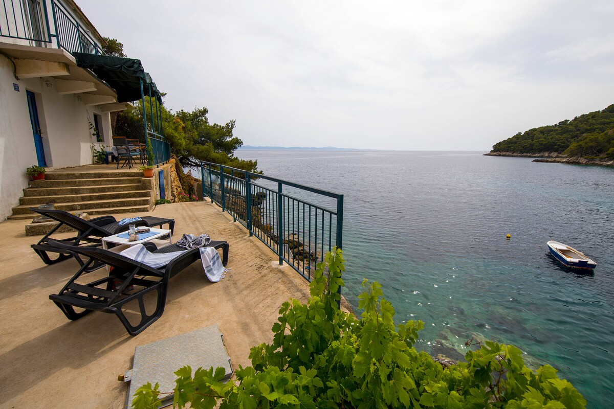 A spacious terrace overlooks the serene bay, featuring sun loungers and a table set for relaxation. The clear waters gently lap against the shore, with a small boat visible nearby. Lush greenery frames the view, creating a peaceful coastal atmosphere.