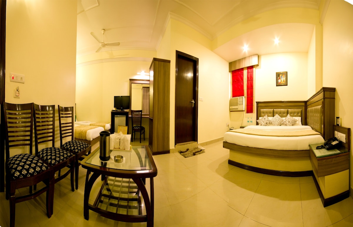 The spacious room is furnished with a large bed, complemented by ambient lighting and wall artwork. A comfortable seating area with four chairs surrounds a coffee table. A TV and air conditioning unit are visible, creating a functional and inviting space.