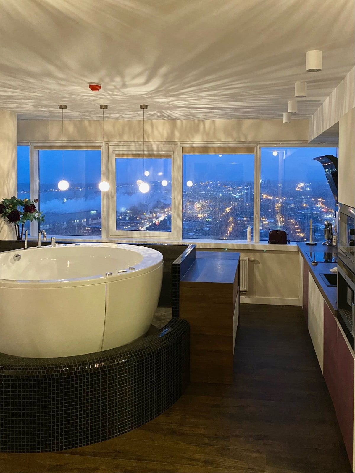 The bathroom features a large round soaking tub set atop a dark tiled platform, with expansive windows showcasing a city view at dusk. Soft pendant lighting illuminates the modern cabinetry and countertops, creating a serene atmosphere.
