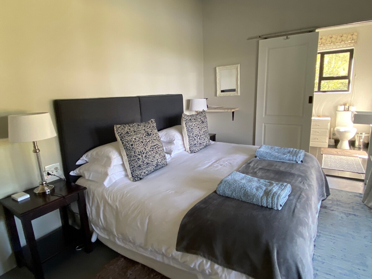 A cozy bedroom is furnished with a spacious bed, adorned with two decorative pillows. A bedside table with lamps flanks either side. A doorway leads to an en-suite bathroom, visible in the background, which features elegant fixtures and a neutral color palette.