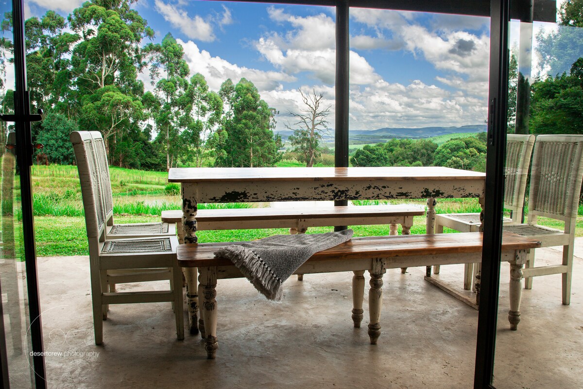 A spacious dining area is showcased with a rustic wooden table and several matching benches. Light fabrics and natural scenery are visible through large glass doors, creating a connection with the green landscape and distant hills beyond.
